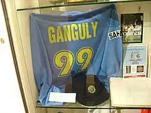 A blue coloured T-shirt displayed at a store window. The T-shirt has the words "Ganguly" and the number 99 below it, both in yellow color. Beside the T-shirt, a picture and an open book is visible.