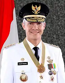 Official portrait of Ganjar Pranowo in 2018, showing him wearing a white ceremonial uniform displaying various medals. He is also wearing a black service cap with a Garuda Indonesia, the national symbol of Indonesia, on it. On the background is the Indonesian flag.