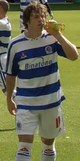 Young white man with dark curly hair wearing blue-and-white sports kit leaves a sports field.