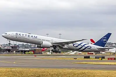 A wide, navy blue plane takes off of Sydney Airport. Qantas airplanes can be seen in the background.