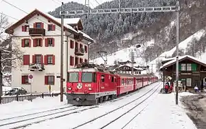 Red train arrives on snow-covered tracks at a two-story building with gabled roof