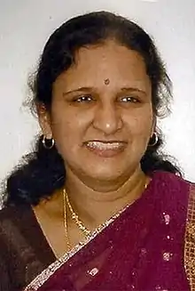 A woman of Indian descent, smiling, seen in portrait view from the bust level up wearing jewelry and a predominantly maroon sari