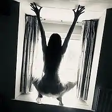 Silhouette of the back of a woman crouched before a window, with hands outstretched above her, pressed against a low ceiling