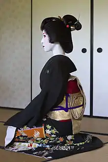 A profile view of a geisha sat kneeling. She wears a black formal kimono, a gold belt, a traditionally styled wig and white make-up with red lips and accents.