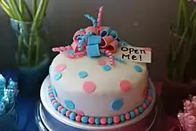 A round cake with a white base color decorated with edible circles and ribbons of both pink and blue coloration. An attached note says "Open Me".