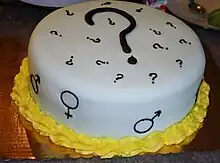 A round white cake with a large black question mark and smaller gender symbols on it.