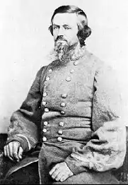 Black and white photo shows a bearded man with long, dark hair. He is seated and wears a gray uniform with two rows of buttons.