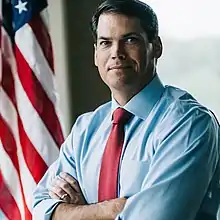 Portrait of Geoff Duncan in blue dress shirt with sleeves rolled up and red necktie, arms folded, in front of a U.S. flag.