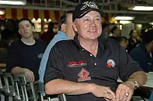 A middle-aged man sitting in a chair and smiling. He is wearing a black T-shirt and baseball cap with sponsors' logos.