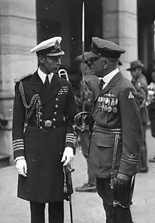 Photograph of two men in formal military uniform. The man on the left is a naval officer's uniform, and has a cap, white gloves, a ceremonial sword, and has medal ribbons on his chest. The man on the right is also in a military officer's uniform and is wearing a cap, leather gloves, and medals. He is carrying a ceremonial sword in his right hand. Other men in military uniforms can be seen in the background.