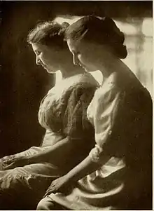 Black and white image of two women with their heads bowed and hands in their lap.  They're both wearing dresses and have their hair pinned back.