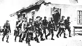 A black and white photograph of a group of a dozen German soldiers marching past a house with weapons slung over their shoulders