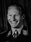 A smiling man wearing a military uniform, and an Iron Cross displayed at the front of his uniform collar.
