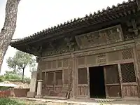 Half a of a red-walled building with large eaves projecting from the corner. A tree trunk is visible on the left-hand side of the image