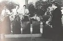 From left to right: three standing saxophone players, one sitting guitar player, one accordion player, one fiddler, one conductor and one barely visible drummer. All are men and wear six-pointed stars.