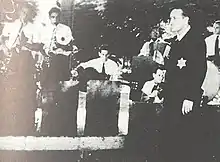 From left to right: two standing saxophone players, one sitting guitar player, one accordion player, one fiddler, one conductor and one drummer. All are men and wear six-pointed stars.