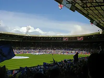 A stadium full of spectators. Those nearest the camera are waving blue and white flags.