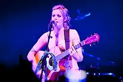 A woman with blonde hair wearing a yellow shirt, playing a guitar and singing into a microphone