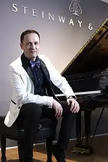 A dark haired man in a tailcoat and a kummerbund, dark bow tie, both hands in the pockets of light-coloured trousers.