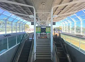 Island railway platform with escalators going down and a metal shelter and glass side walls