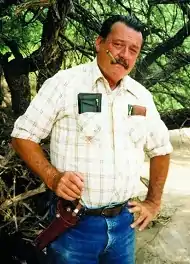 Glenn Boyer standing smoking a cigar with a holstered six-shooter on his hip.