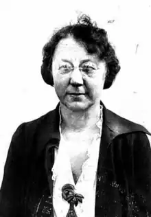 A middle-aged white woman with short dark hair, wearing a dark jacket over a white blouse, and glasses