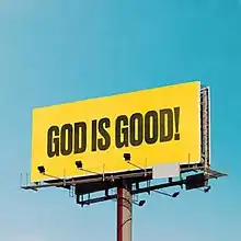 The album cover is a billboard with the text "God Is Good!" capitalized in a black sans serif font against a yellow backdrop. The background of the billboard is a clear blue sky.
