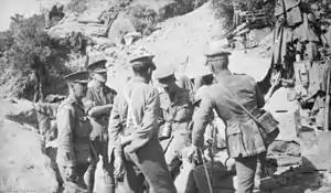 Six men in uniform with peaked caps satand and sit in a circle talking to each other. In the background are sandbags, tins and clothing hung out to dry.