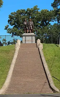 Photograph showing a long, broad flight of steps leading to a large metal statue of two men. There are about 50 steps. The two men in the statue are roughly life-sized, and are standing on a large stone pedestal. The pedestal has bronze plaque that reads "Goethe und Schiller".