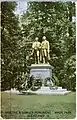 Picture postcard of a large bronze statue of two men on top of a stone pedestal. There are several wreaths that have been placed at the base of the pedestal. The statue is standing in a grassy clearing in front of trees. There is printing along the bottom of the postcard that says "Goethe and Schiller Monument, Wade Park, Cleveland, O."