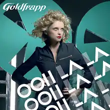 A blonde-haired woman at the centre standing with her hands on her hips and looking off to the right of the image. Text overlays the woman and the bottom right of the image saying "Ooh La La" three times. Text in the upper left corner says "Goldfrapp". In the background is a black wall along with two teal-coloured circles with triangle-shaped holes cut out of them.