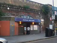 An entrance under a railway brick viaduct with a blue sign reading "GOLDHAWK ROAD STATION" in white letters and two women walking in front all under a grey sky
