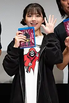 Miho Watanabe holding a Blu-Ray case and waving toward camera