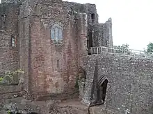 A castle, with a flat fronted tower facing the viewer with a stained glass window in the middle of it; a stone causeway is on the right of the picture, leading to a gateway to the right of the tower – a partially filled arch is supporting the causeway.