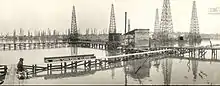 A black-and-white photograph showing a field of oil derricks and thin wooden boardwalks built over the water.