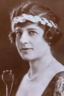A white woman with short dark wavy hair, wearing a headband with a laurel-crown effect, and a dark dress with a deep scooped neckline, and a strand of pearls