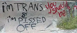 Graffiti on a concrete wall, in red and black. The black graffiti reads, "I'm trans & I'm pissed off". The red graffiti reads, "You should be".