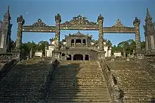 Staircase leading to a building of dark stone. A simple decorated gate is at the top of the staircase.