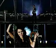 The top of the image shows a man with gelled, black hair, a white T-shirt, jeans and a black leather jacket that stands behind a swing at nighttime. The bottom of the image shows a female teenager with long, brown hair facing forward as she grasps chains with both of her hands.