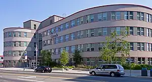 A four-storey stone building with a curved front, broad on the right and narrow on the left, seen from across a street. Its windows are set in darker bands of stone and it has red trim. The first storey is faced in rusticated stone; the ones above are smooth. On the front is the word "Canada" with a miniature Canadian flag on the right, between the third and fourth storeys. In front small deciduous trees are planted, and several flags including the Canadian flag fly from poles in front of the main entrance near the junction of the two curves. Two cars are parked along the street in front.