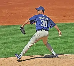 A man wearing a blue cap, blue top, and light-colored trousers, holding a baseball. The back of his shirt reads "Maddox 30"