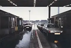 View of two lines of vehicles passing between two buildings, with four passport control booths visible, under a corrugated metal roof. A long line of vehicles stretches into the distance below towers ringed with searchlights.