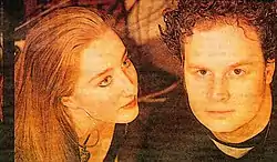 Head shot of two people. At left is a red-brown haired woman looking to her left. She wears a very large circular earring in her right ear lobe. At right is a dark, curly-haired man who is staring forward. He wears a dark top and has side-burns.