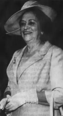 A smiling older Mexican woman wearing a light-colored suit and matching wide-brimmed hat, and gloves