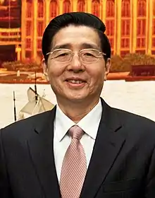 a smiling, clearly aged man with wavy haircut, wearing glasses, a white shirt, a suit and a light red tie
