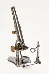 A stand microscope created by Joseph Gutteridge, a weaver from Coventry. The microscope allows coarse and fine adjustments, and its height is adjustable by a set screw. It consists of two objectives each with a screw on cap and a slidecarrier that is moveable laterally. The mirror also has a cap. It has a bench magnifier, lens moveable through 360° and has a base weighted with lead.