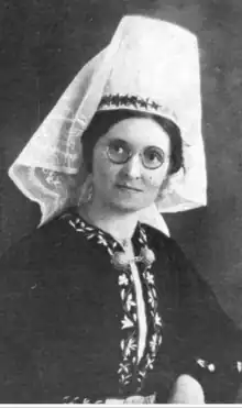 A young white woman with dark hair, wearing round glasses and a tall white fabric headpiece and dark embroidered jacket.