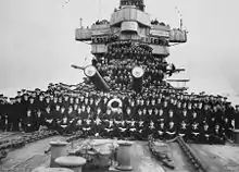 A large number of men posing for a photo on the foredeck of a warship. Two of the ship's gun barrels are visible in the middle of the group.