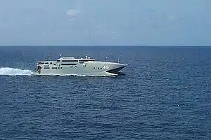 A warship-grey catamaran travelling at speed (from right to left) on the open sea