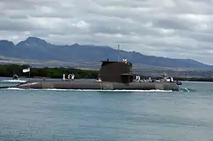 Side view of submarine with sailors on deck, at sea in front of a hilly coast